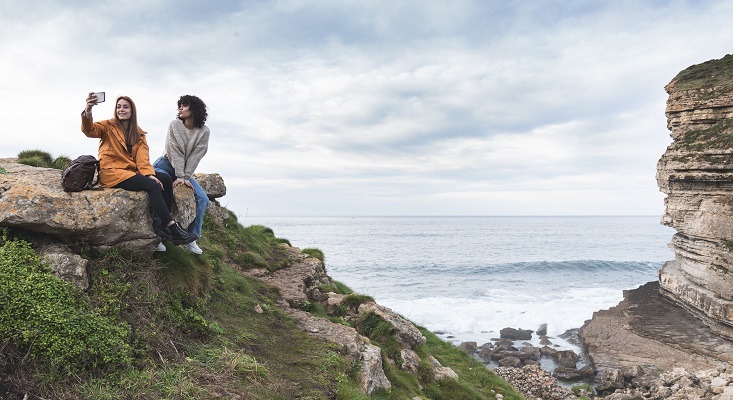 Female friends taking selfie while sitting on cliff by sea at Acantilado El Bolao